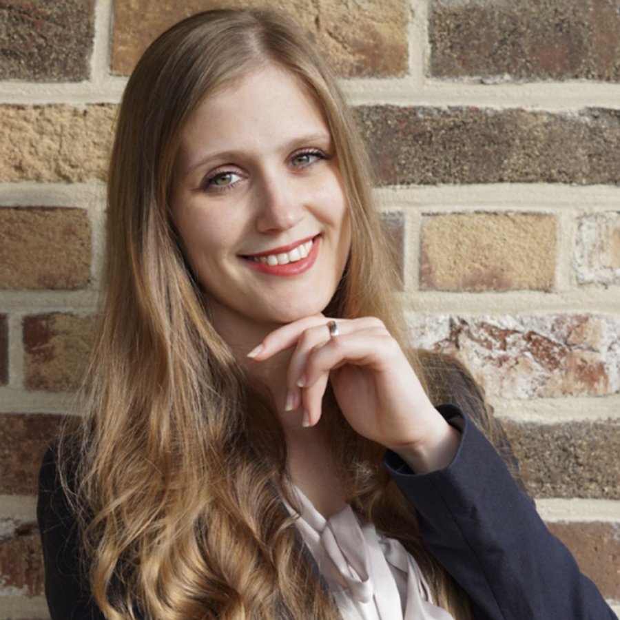 A smiling woman with long blonde hair, red lipstick, and a blazer, resting her chin on her hand, against a brick wall.