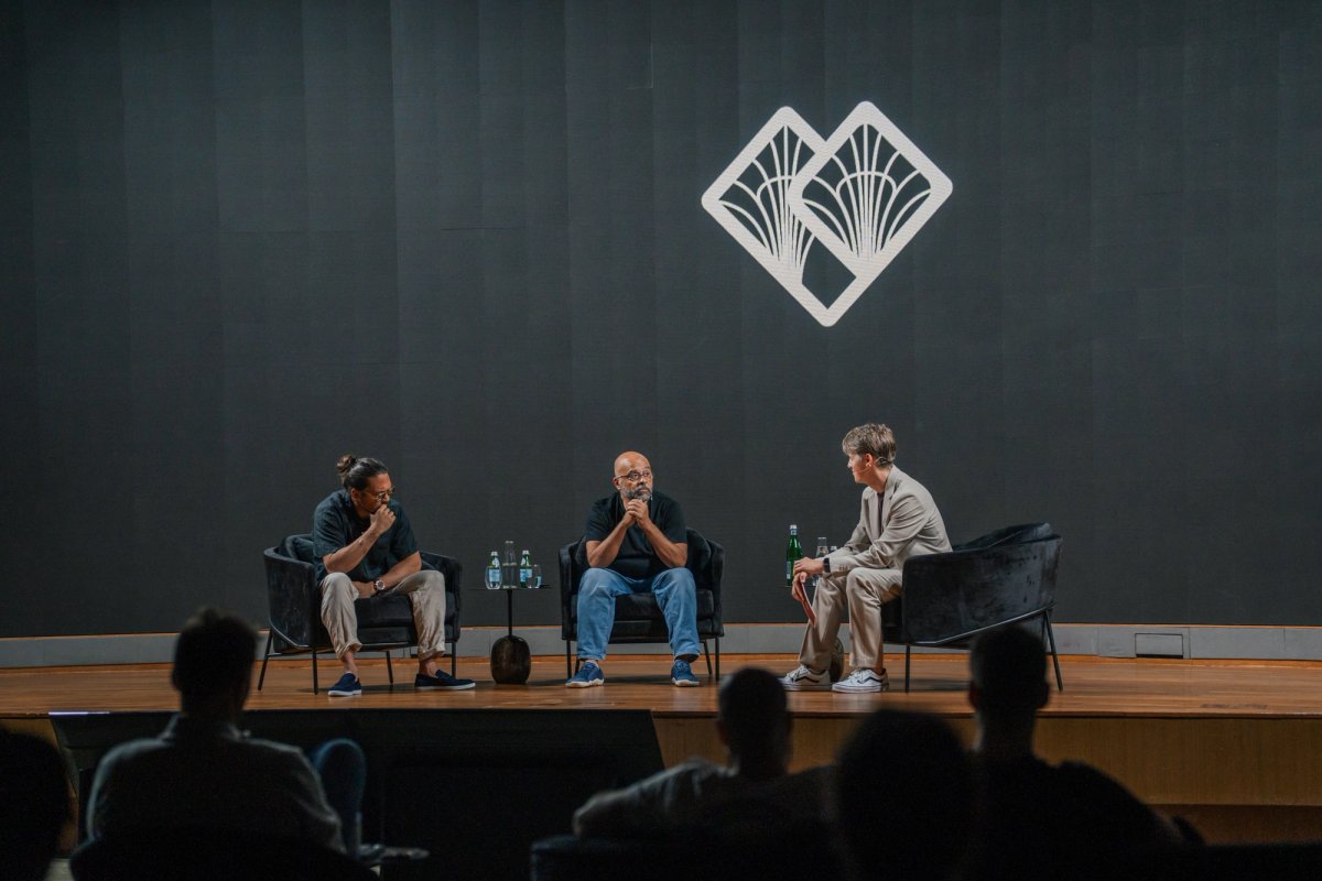 Three people sit on a stage for a panel discussion, with a white logo on the dark wall behind them.
