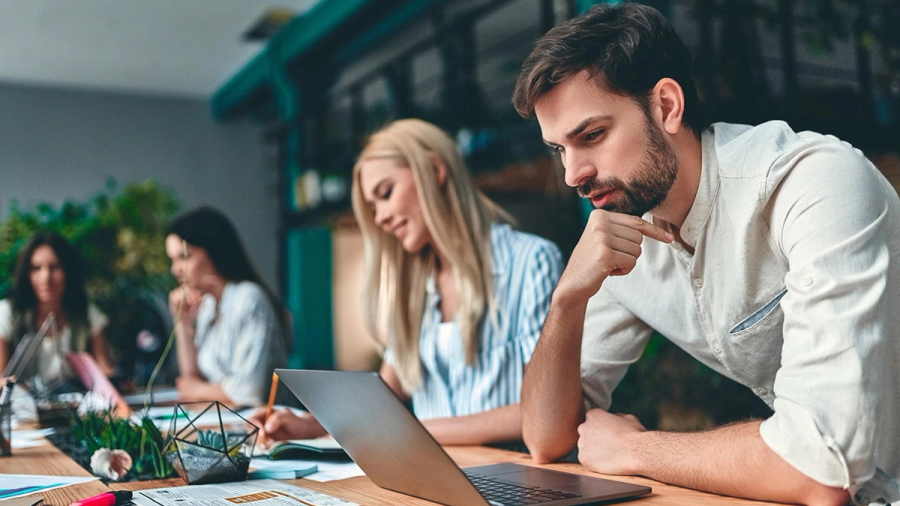 A man intently focused on a laptop while colleagues work at a table.