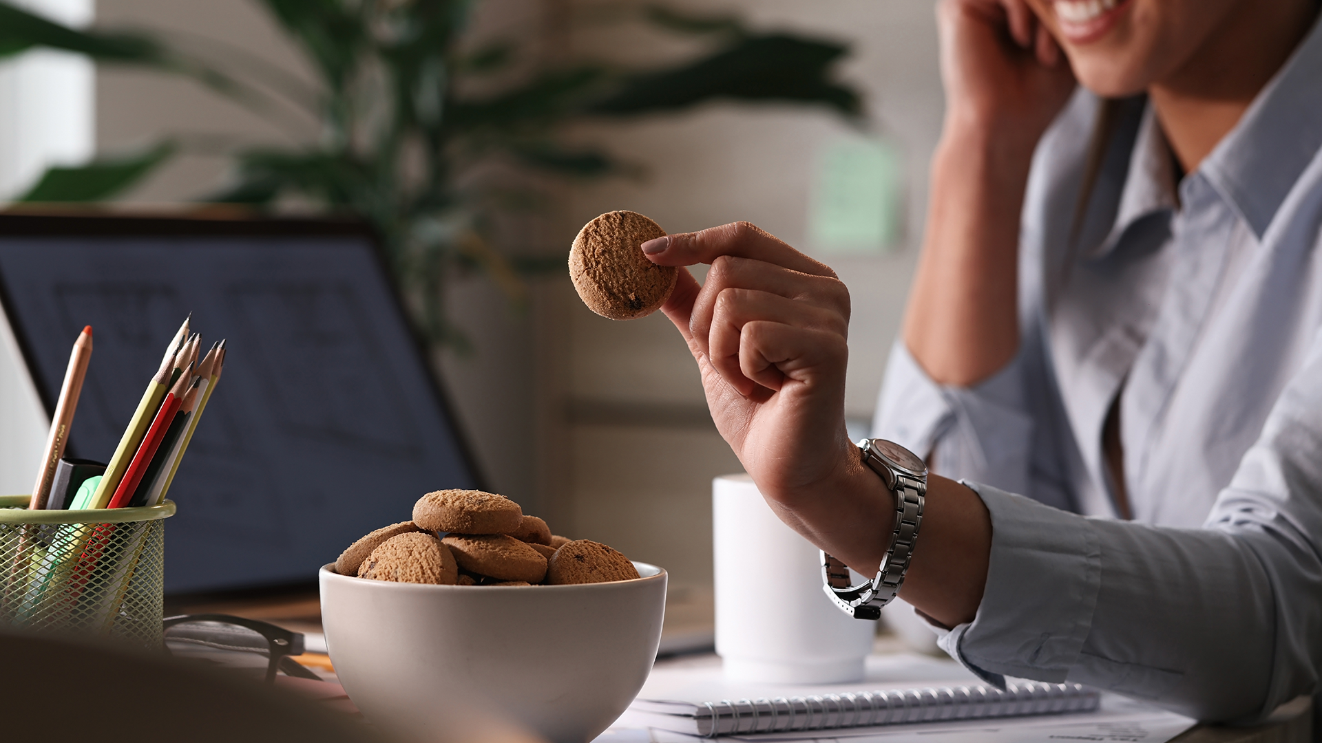 A person holding a cookie at a desk with a bowl of cookies and a laptop.
