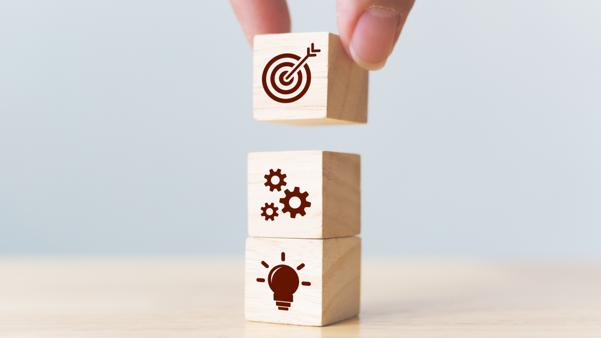 Hand stacking wooden blocks with icons for ideas (lightbulb), process (gears), and goals (target).