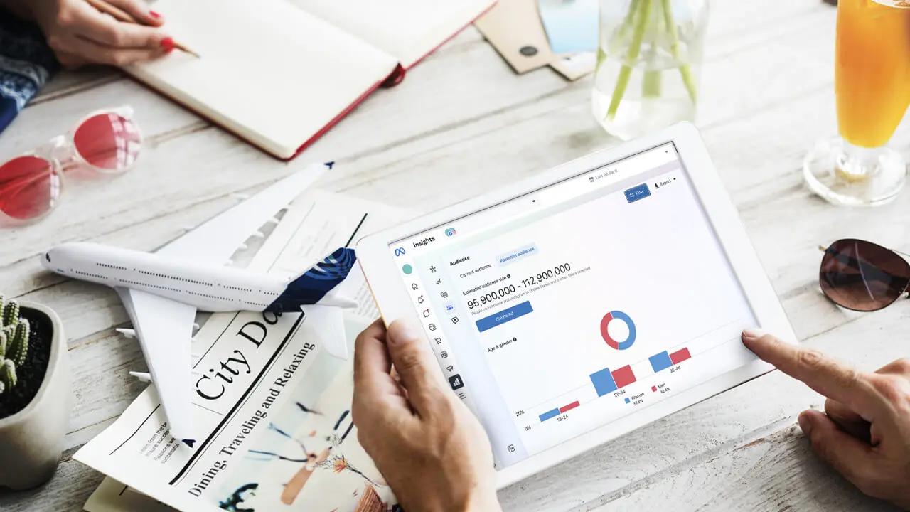 Two people at a table, one using a tablet displaying data, surrounded by a model airplane, travel magazine, and sunglasses.