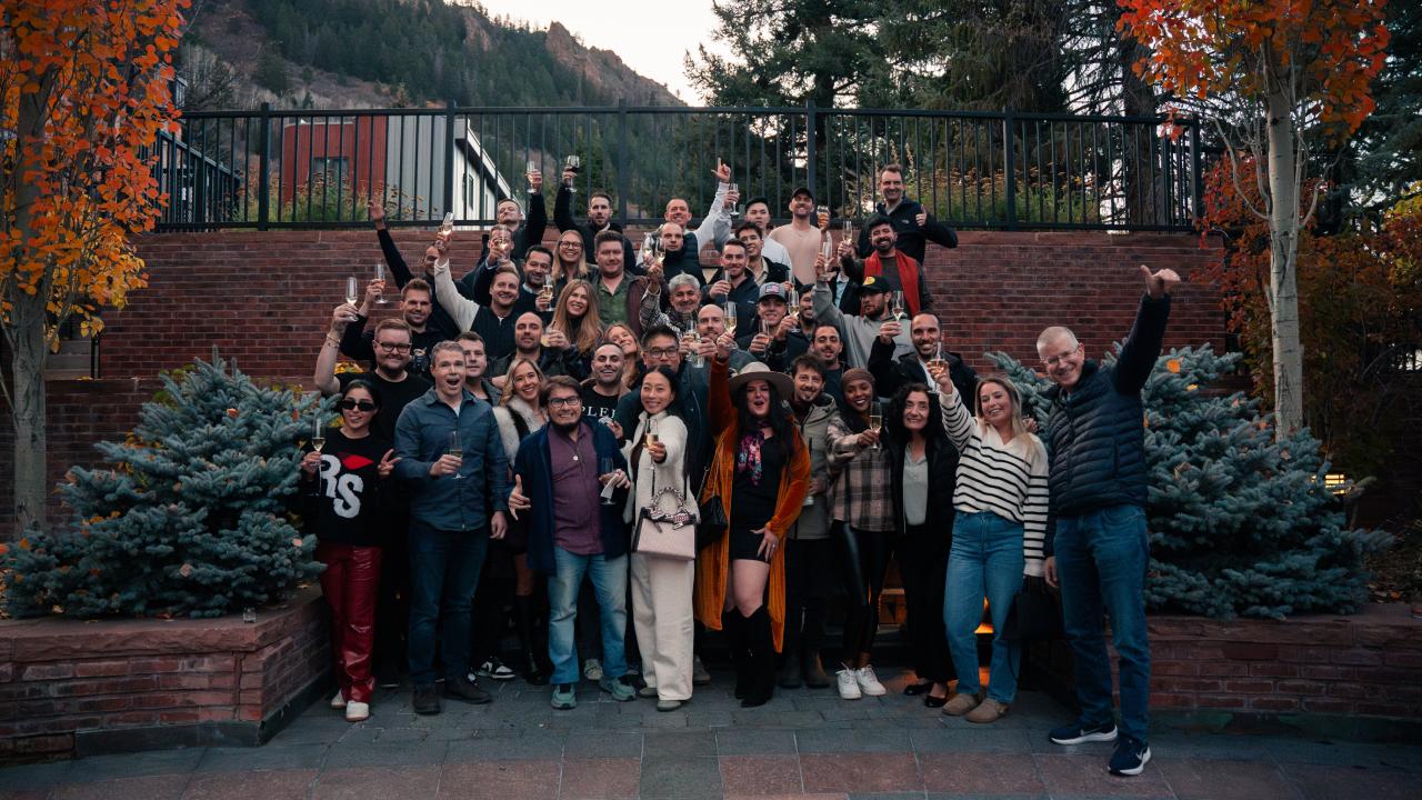 A large group of people cheering with raised glasses on outdoor steps against an autumn mountain landscape.