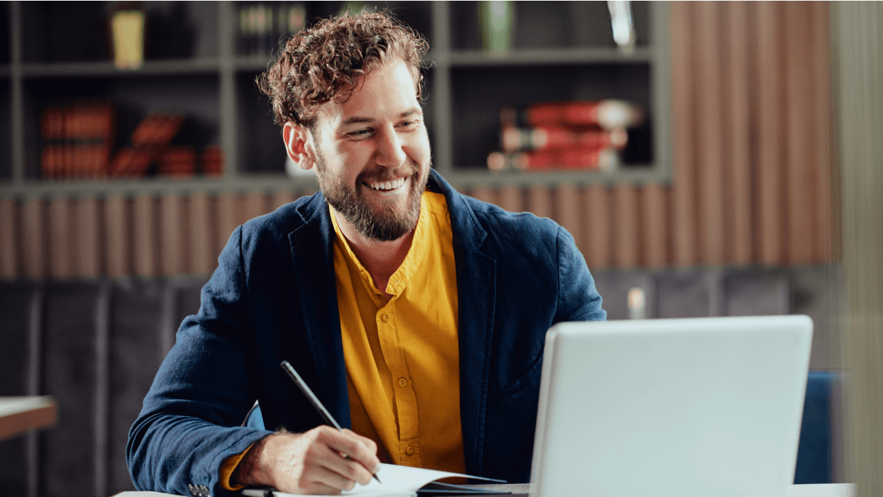 A smiling bearded man writing in a notebook at a desk with a laptop.