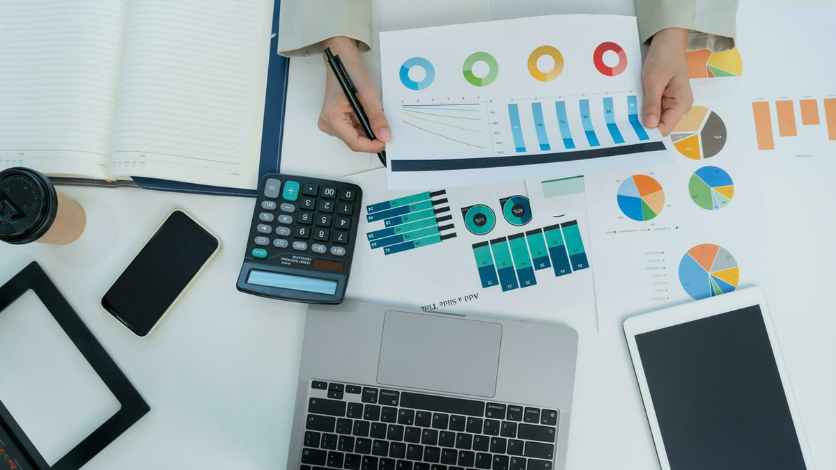A person's hands analyze data charts on a desk with a laptop, calculator, phone, and tablet.