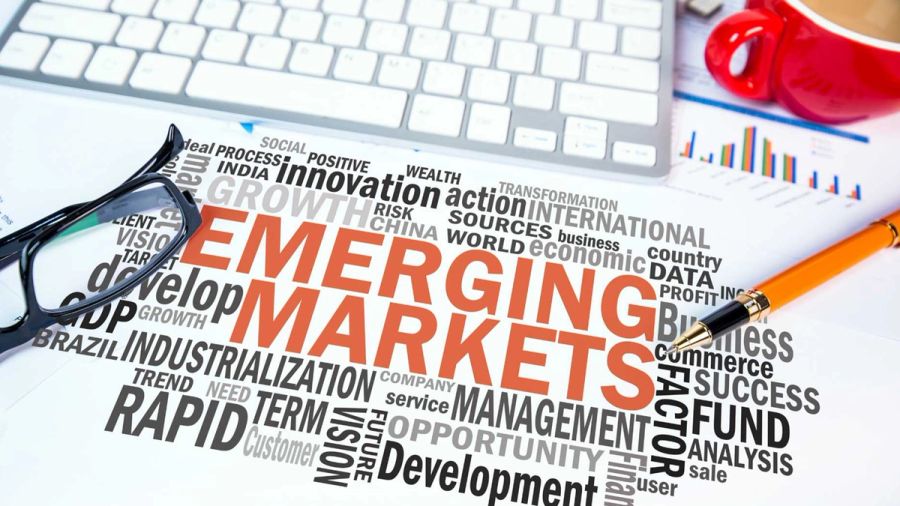 Word cloud centered on "EMERGING MARKETS" with related economic terms, on a desk with a pen and glasses.