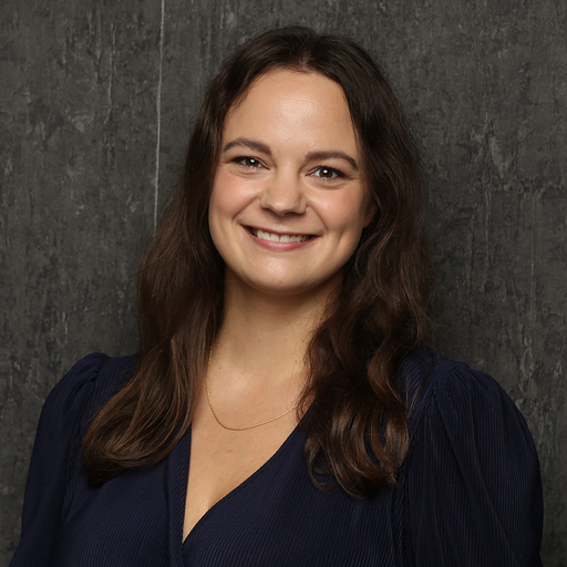 A smiling woman with long brown hair in a dark blue dress and a gold necklace against a dark background.