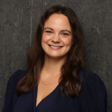 A smiling woman with long brown hair in a dark blue dress and a gold necklace against a dark background.
