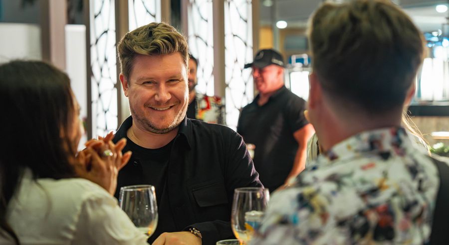 A man with light brown hair smiles at a woman across a table with wine glasses.