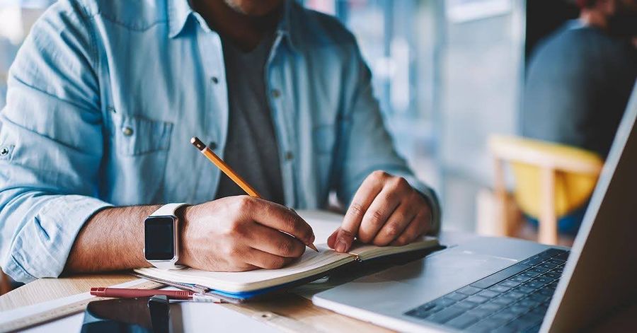 Person wearing a smartwatch writing in a notebook with a pencil, a laptop open on the desk.