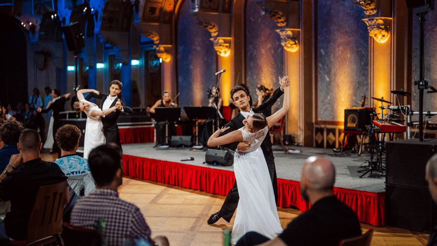 Two couples ballroom dancing on a polished floor in an elegant hall with a live band and an audience watching.