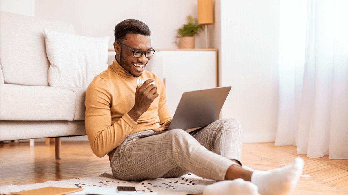 Smiling man sitting on the floor with a laptop and a cup.