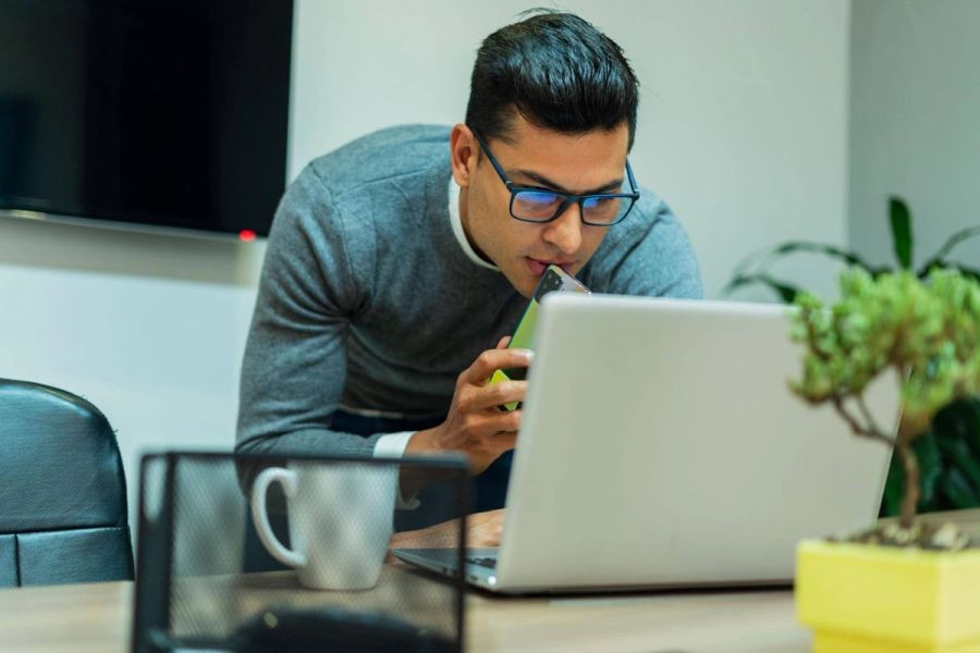 Man in glasses speaking into a smartphone while leaning over a laptop.