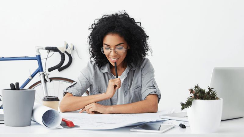 A thoughtful Black woman with curly hair and glasses works on blueprints at a desk, with a bicycle in the background.
