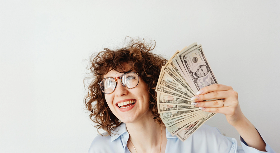 A smiling woman with curly hair and glasses holds a fan of dollar bills.