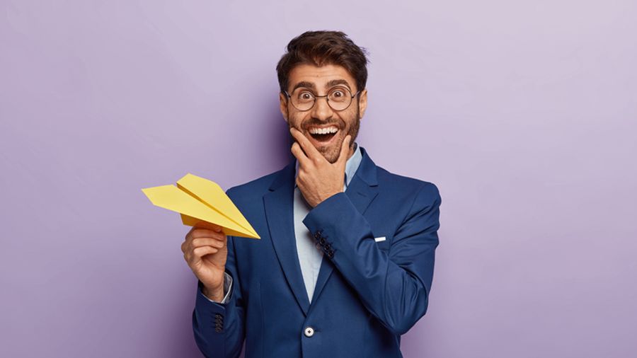 An excited man in a suit and glasses holds a yellow paper airplane.