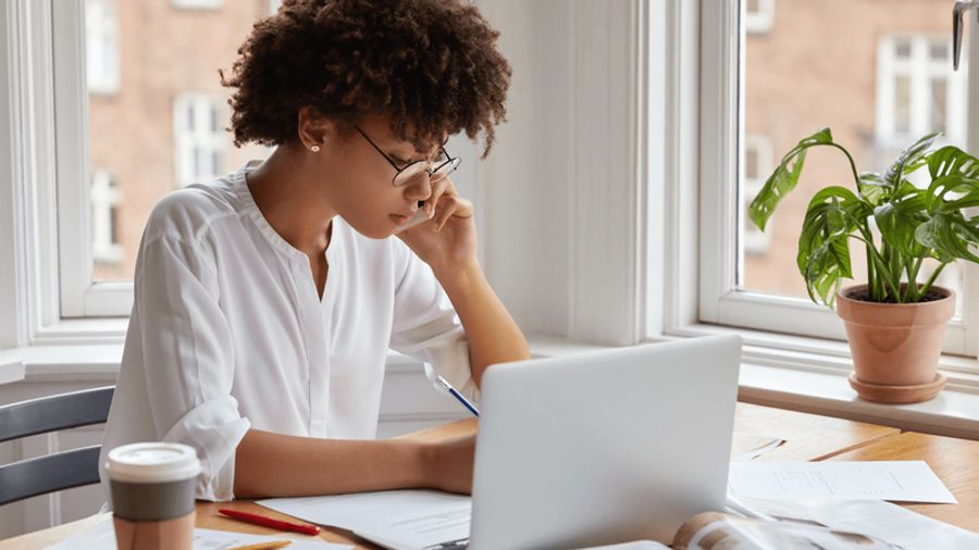 A woman with glasses writes at a desk with a laptop, talking on a phone by a window.