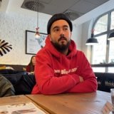 A bearded man in a black beanie and red hoodie sits with crossed arms at a wooden table in a cafe, looking up and right.