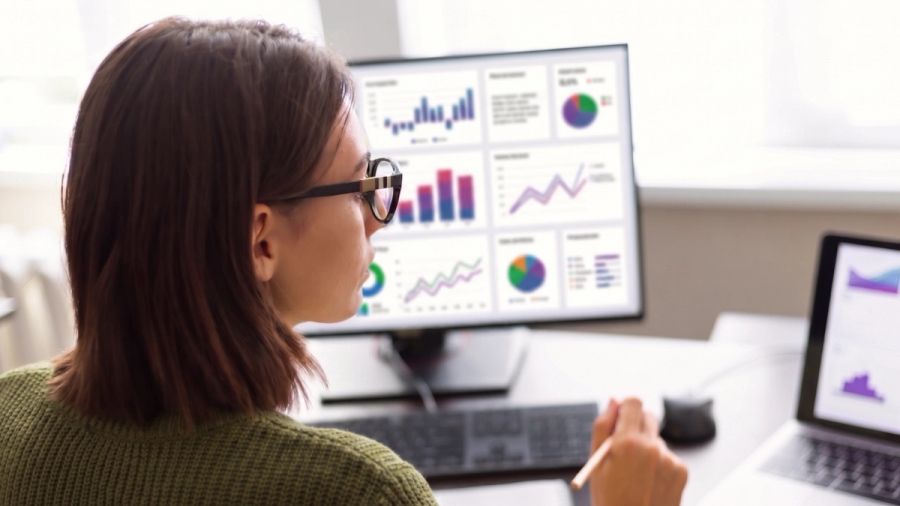 A woman in glasses views a computer monitor displaying a data dashboard with charts and graphs.
