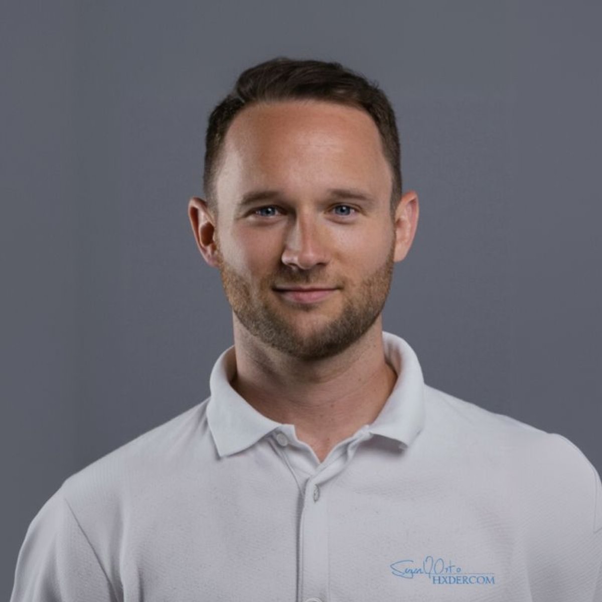 A smiling man with short brown hair and a beard, wearing a light polo shirt against a gray background.