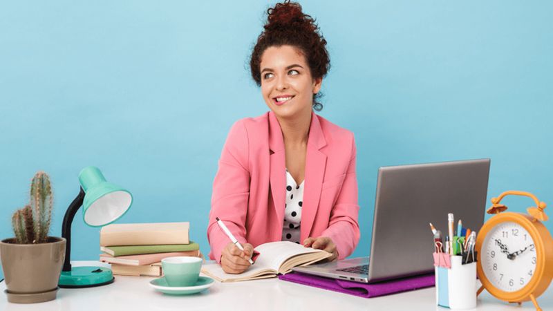 A smiling woman in a pink blazer writes in a notebook at a desk with a laptop.