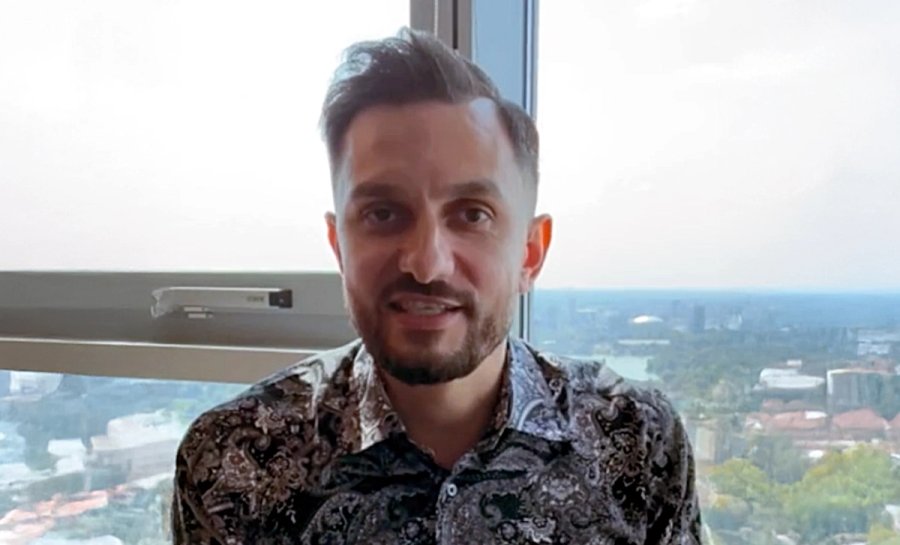 Homem com barba e camisa estampada sorri em frente a uma paisagem urbana vista por uma janela.