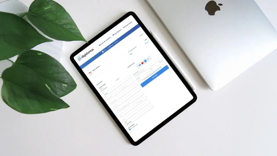 Tablet displaying a Digistore24 payment form, next to a silver laptop and a green plant on a white desk.