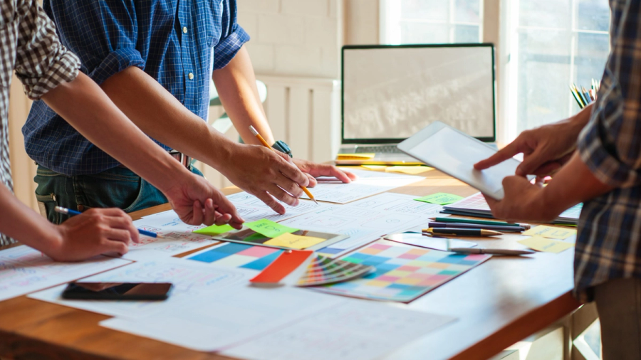 Three people collaborating on design documents, color palettes, and a tablet on a desk.