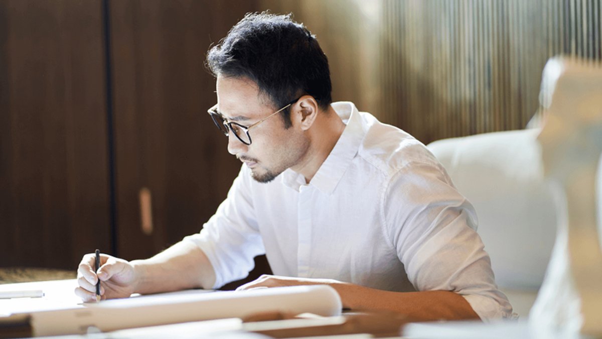 A man in a white shirt and glasses draws on paper.