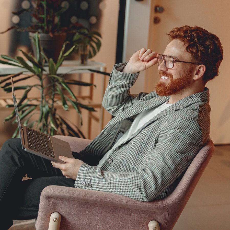 A smiling man with red hair and a beard, wearing a blazer, adjusts his glasses while holding a laptop.
