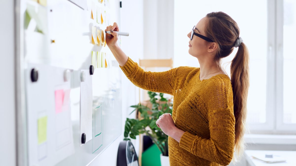 A woman in glasses and a yellow sweater writing on a whiteboard with sticky notes.
