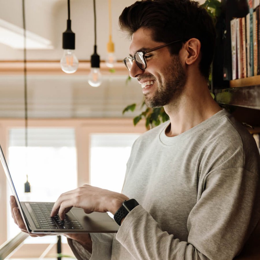 A smiling man in glasses and a grey shirt stands, typing on a laptop.