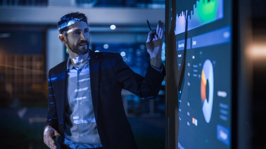 A bearded man in a suit points to a large digital screen displaying data charts and graphs in a dark, blue-lit room.