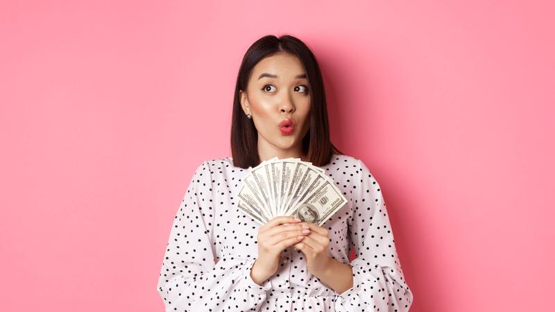 A surprised woman holding a fan of dollar bills against a pink background.