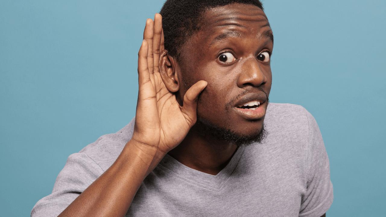 A Black man cups his hand behind his ear, leaning in with wide eyes and an open mouth, as if listening intently.