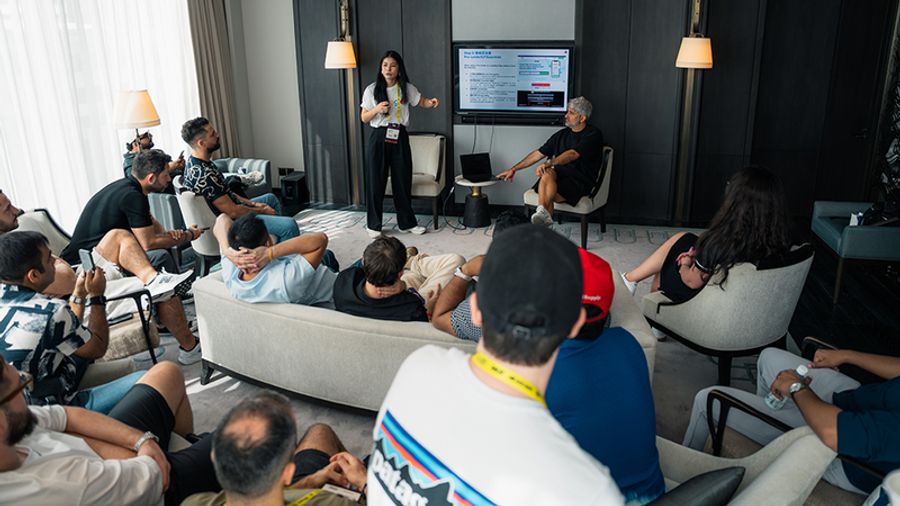 A woman presents with a microphone to a casually seated audience in a modern room, while a man points to a presentation on a large screen.