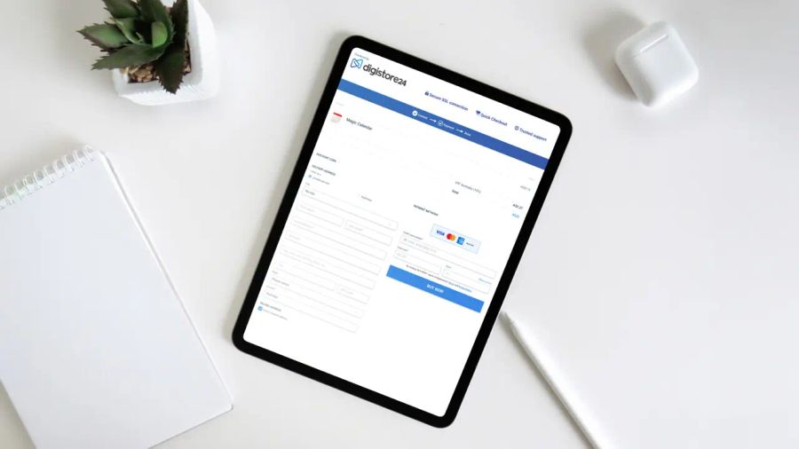 A tablet displaying a Digistore24 checkout page on a white desk with a notebook, plant, stylus, and wireless earbuds.
