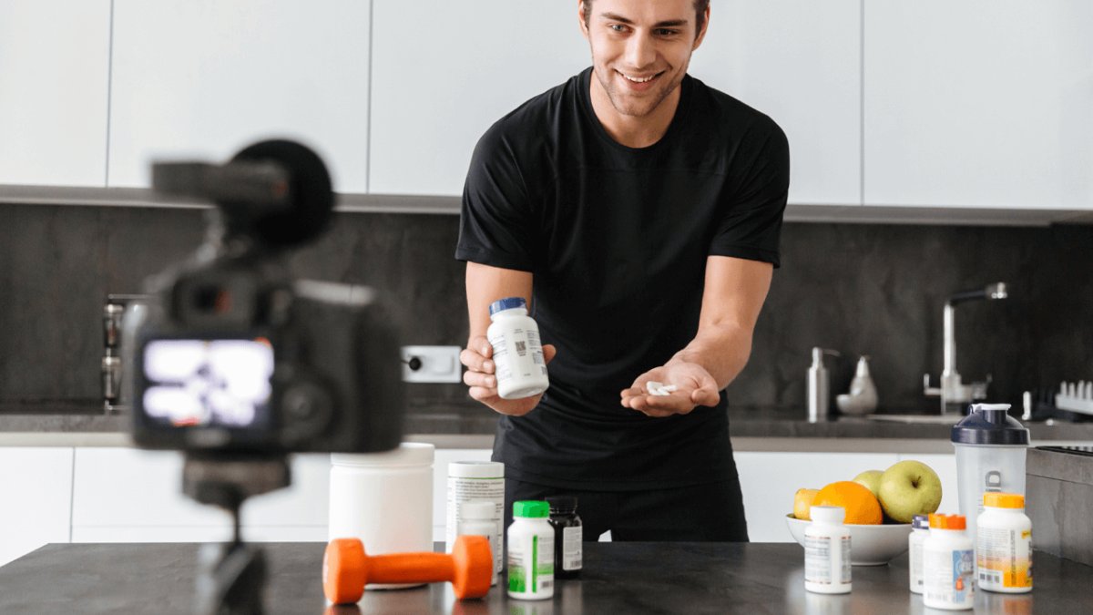 A smiling man filming a video in a kitchen, holding a supplement bottle and pills.
