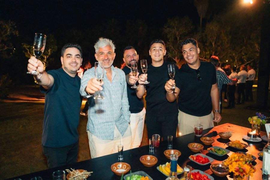 Five smiling men raise champagne glasses in a toast at an outdoor night event.