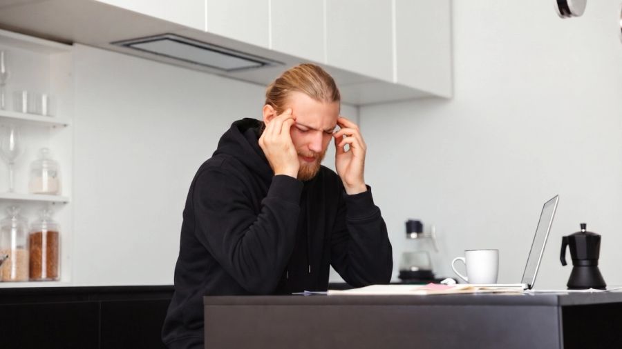 Stressed man with hands on temples at a kitchen counter with a laptop.