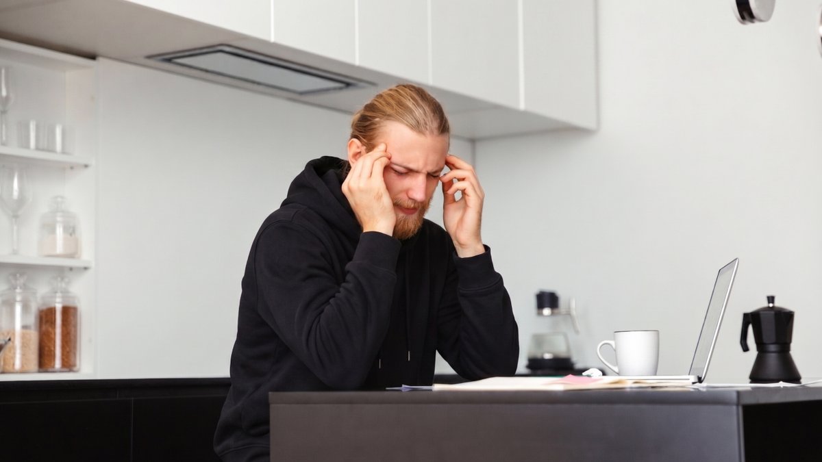Stressed man with hands on temples at a kitchen counter with a laptop.