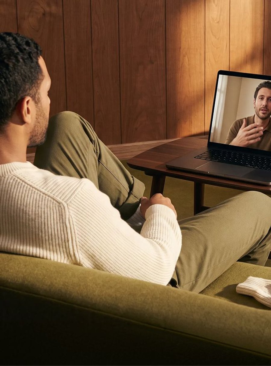 Man on a green couch watching a video call on a laptop with another man onscreen.