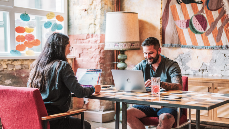 A man and woman work on laptops at a table in a coworking space.