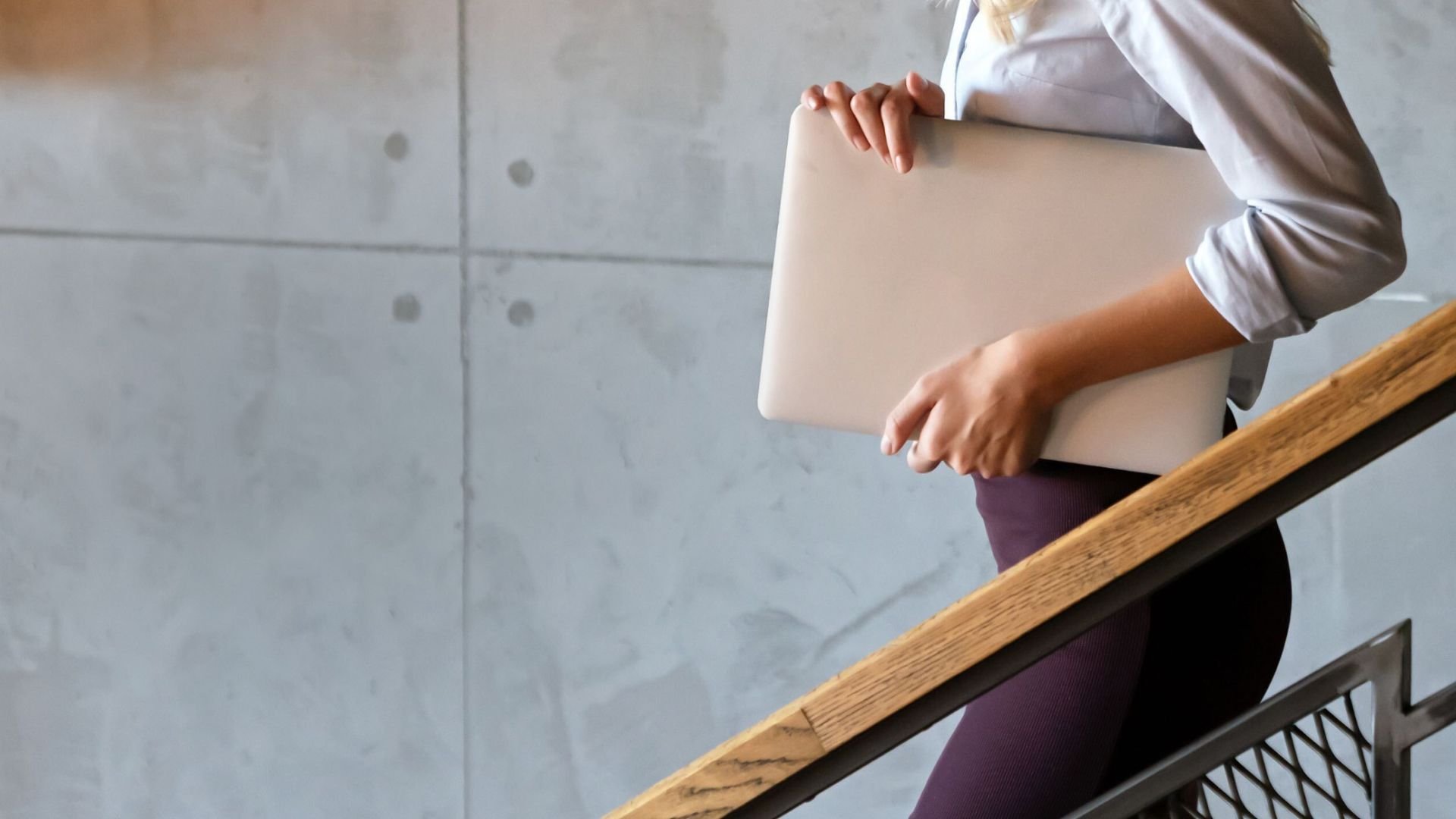 A woman walks up a modern staircase, holding a laptop.