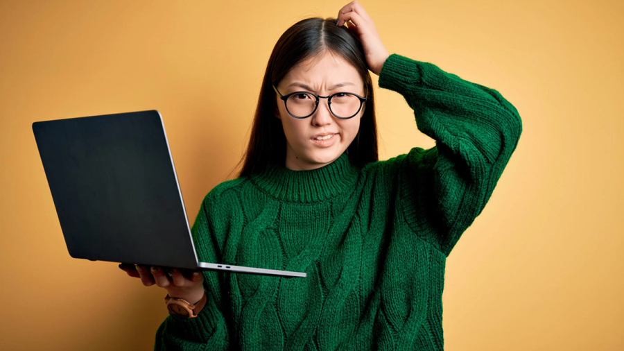 A confused young Asian woman in a green sweater and glasses holds a laptop and scratches her head.