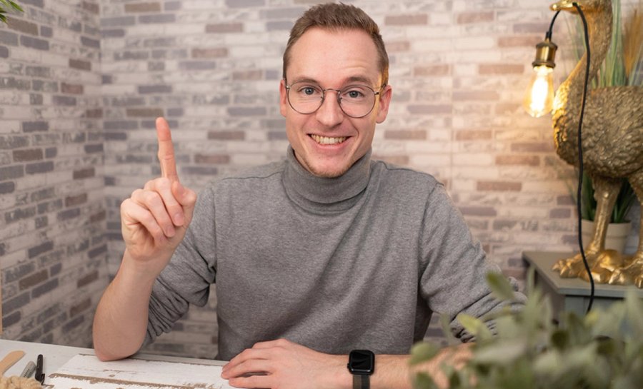 Smiling man in glasses and a grey turtleneck points his index finger upwards.