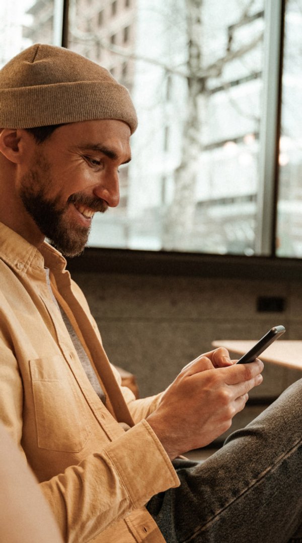Un homme souriant portant un bonnet et une chemise jaune regarde son téléphone.