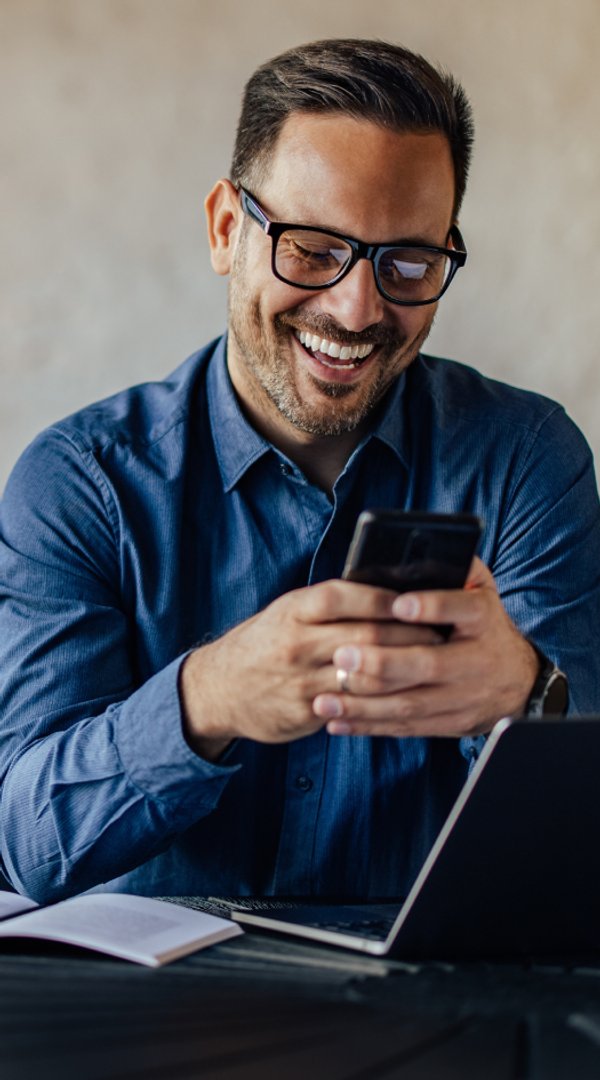 Un hombre sonriente con camisa azul y gafas mira su smartphone.
