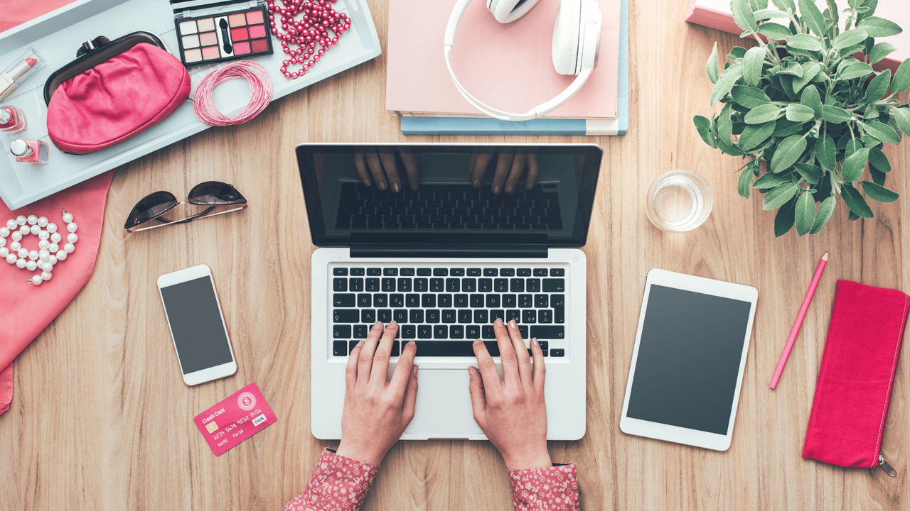 Hands typing on a laptop surrounded by a smartphone, credit card, tablet, and pink personal items on a wooden desk.