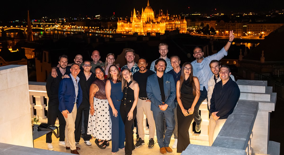 A diverse group of people smiling on a rooftop at night with the illuminated Budapest Parliament, river, and city skyline in the background.